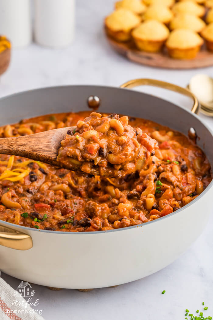 chili mac and cheese in a skillet with a wooden spoon holding up some of the mixture, with cornbread muffins in the background