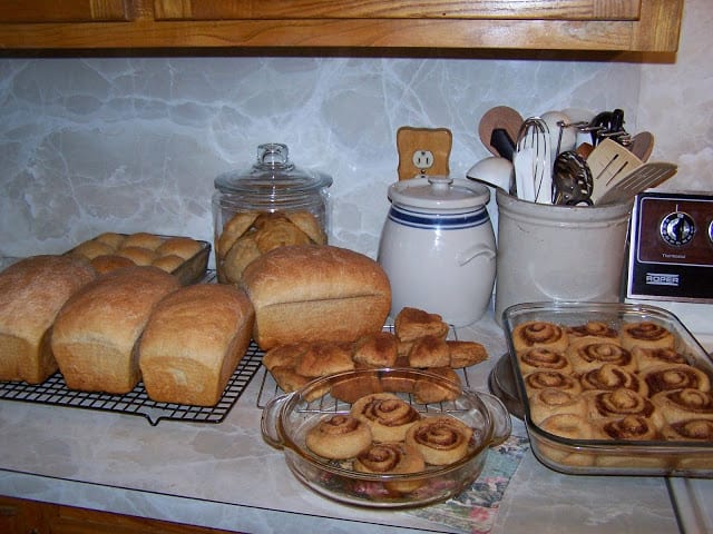 bread baking for busy moms: four loaves of whole wheat bread, 1 square pan with biscuits, two glass pans with cinnamon rolls, and a stack of scones on a cooling rack