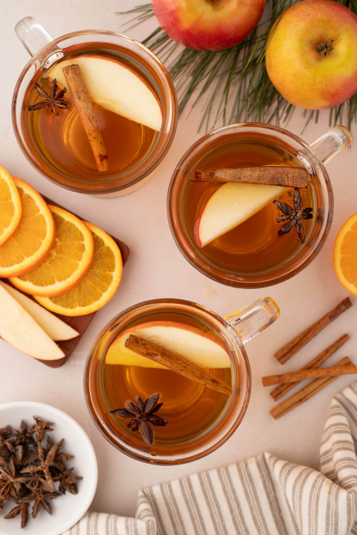Three clear glass mugs filled with homemade apple cider and apple slices and cinnamon sticks.