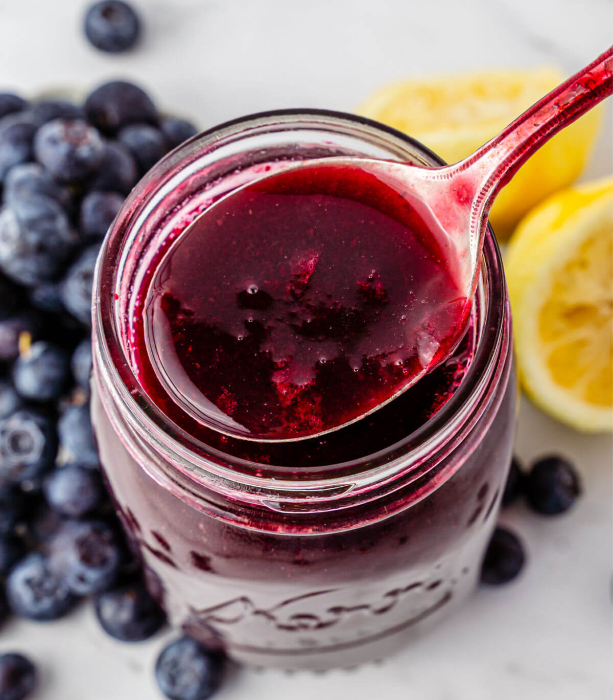 Homemade blueberry syrup in a mason jar next to a pile of blueberries and some lemons.