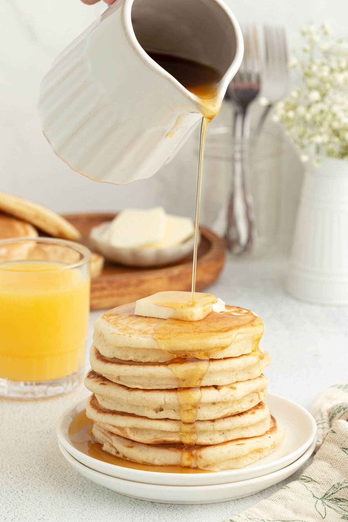 A pitcher of maple syrup pouring syrup onto a stack of sourdough pancakes.