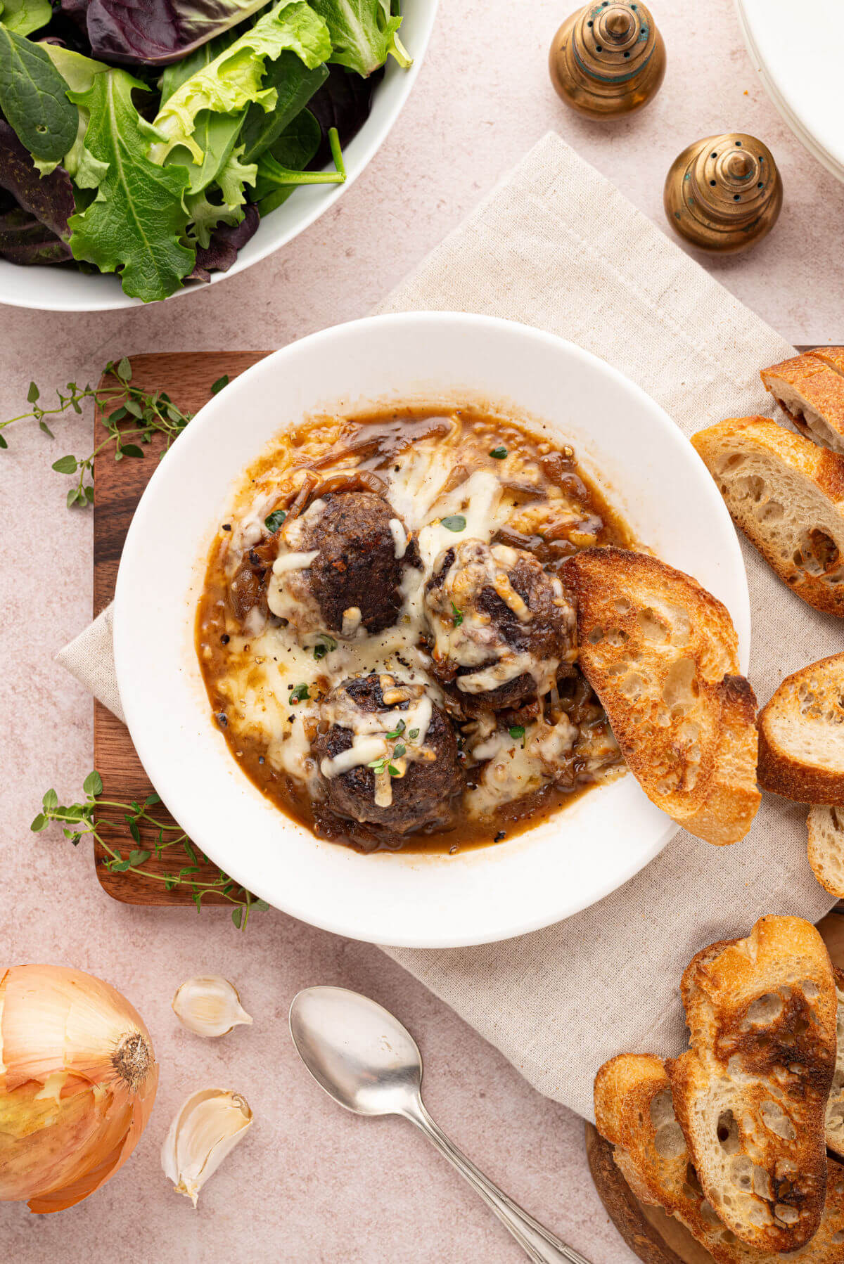 French onion meatballs with melted cheese in a serving bowl with French bread slices and salad.
