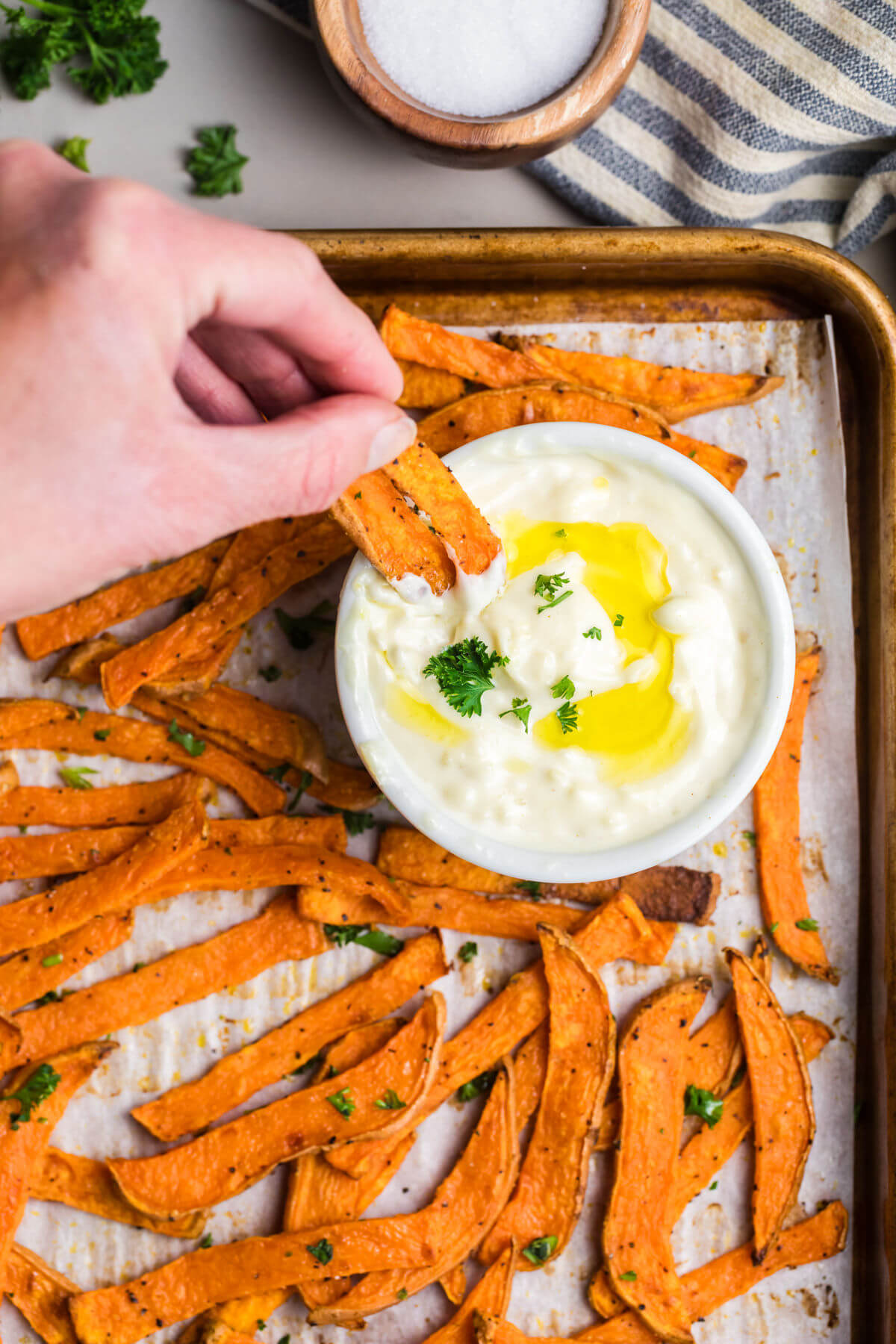 Crispy sweet potato fries on a baking sheet with garlic aioli sauce and a hand dipping fries in the sauce.