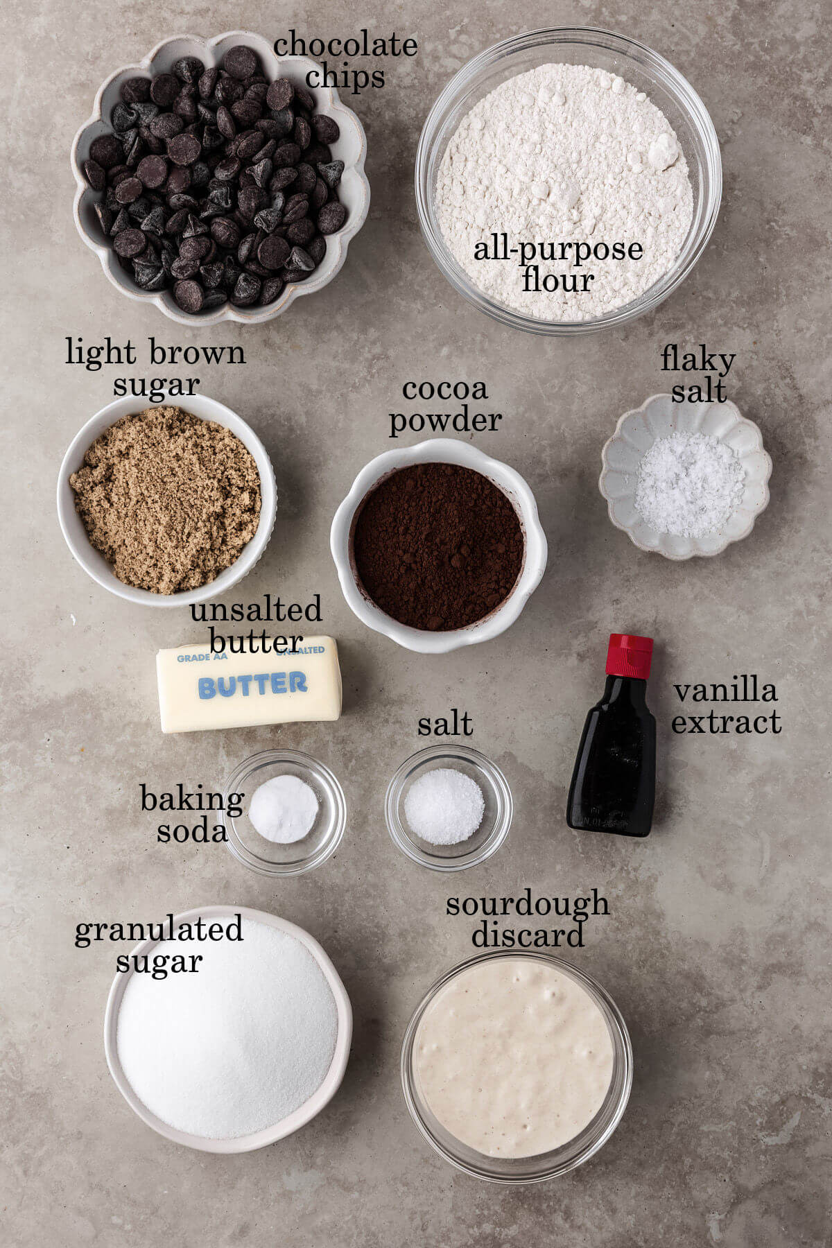 Ingredients for sourdough chocolate cookies in small bowls with text overlay labeling each ingredient.
