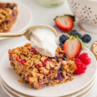 Slice of baked oatmeal with berries on a plate with a spoon adding whipped cream to the top.