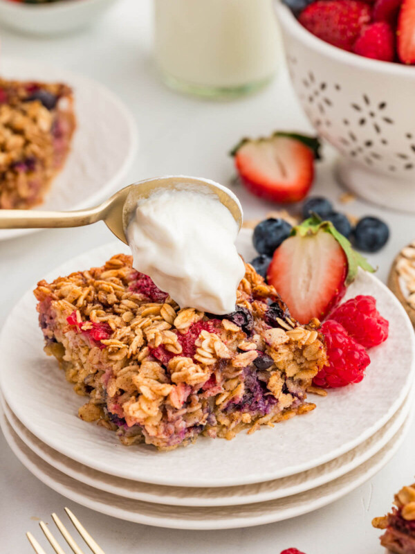 Slice of baked oatmeal with berries on a plate with a spoon adding whipped cream to the top.