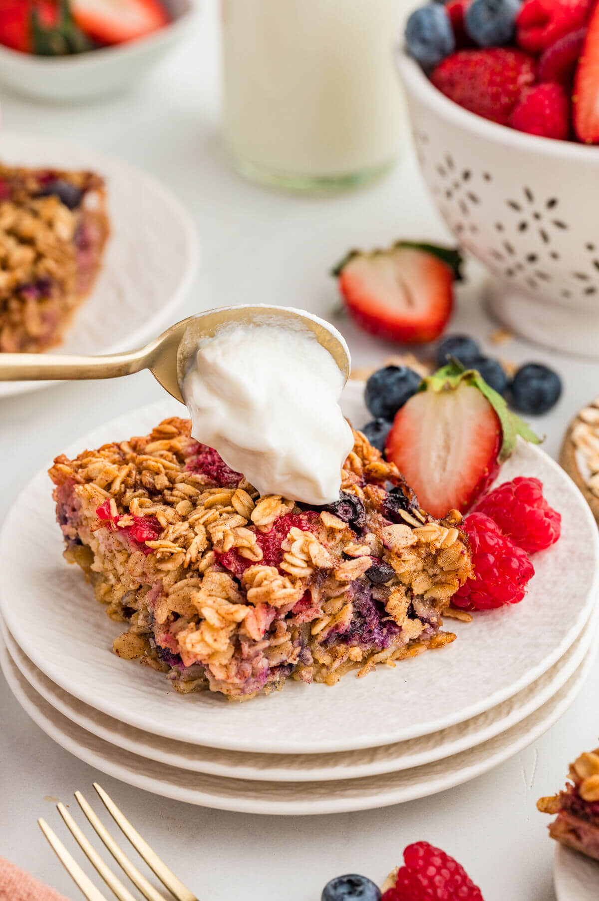 Slice of baked oatmeal with berries on a plate with a spoon adding whipped cream to the top.