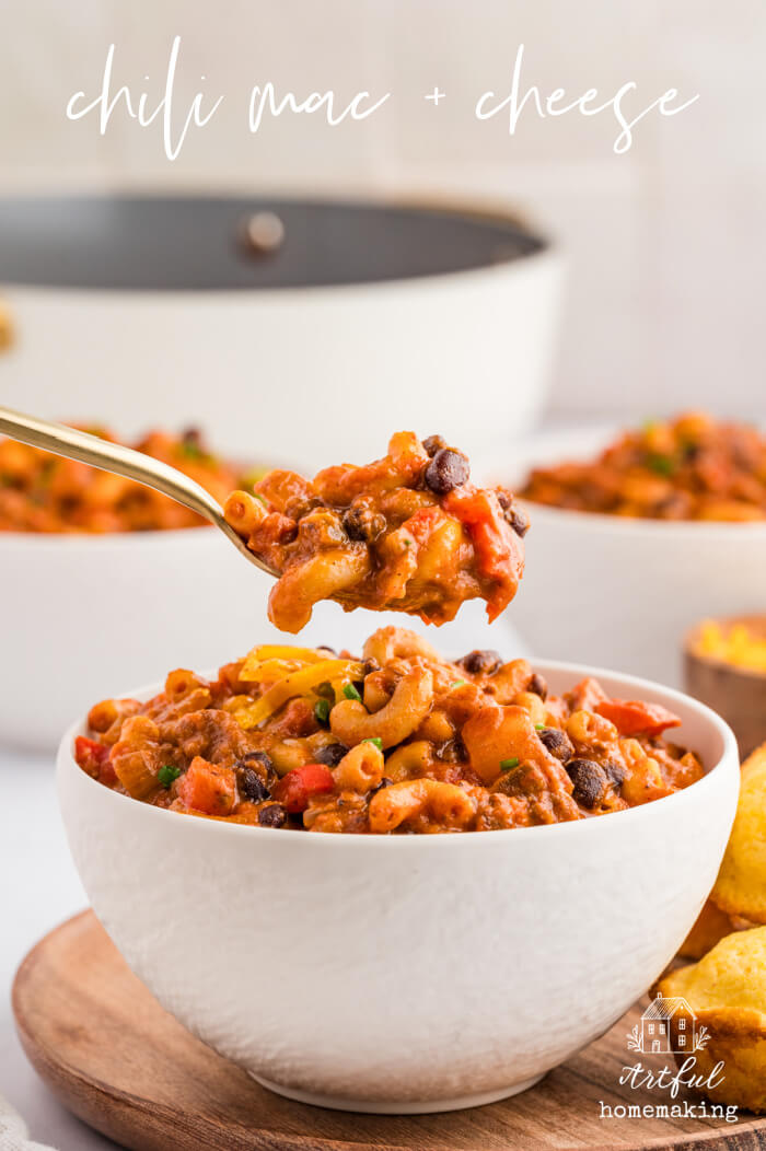 chili mac in a white bowl on a wooden plate with a gold spoon holding up some of the chili mac