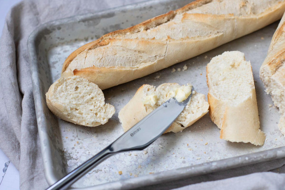 Homemade french baguettes on a baking sheet with a butter knife spreading butter on them.