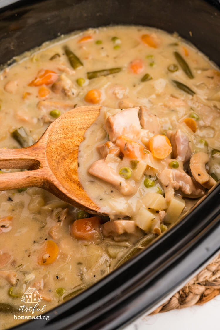 a slow cooker full of chicken stew with a wooden spoon holding up some of the stew