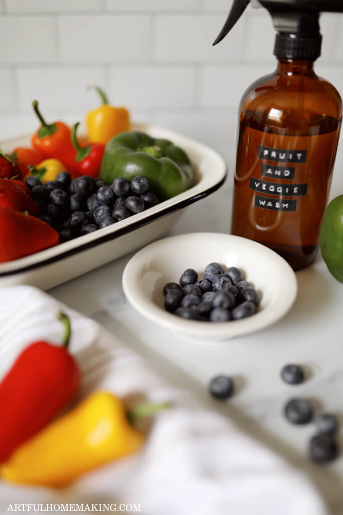 blueberries in a small white bowl, a dish of colorful fruits and vegetables, and a spray bottle on a kitchen counter