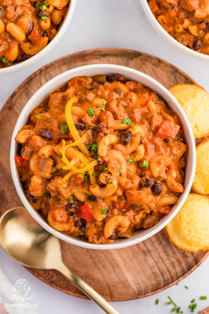 three white bowls with chili mac on a table, with cornbread muffins next to the bowl in the foreground
