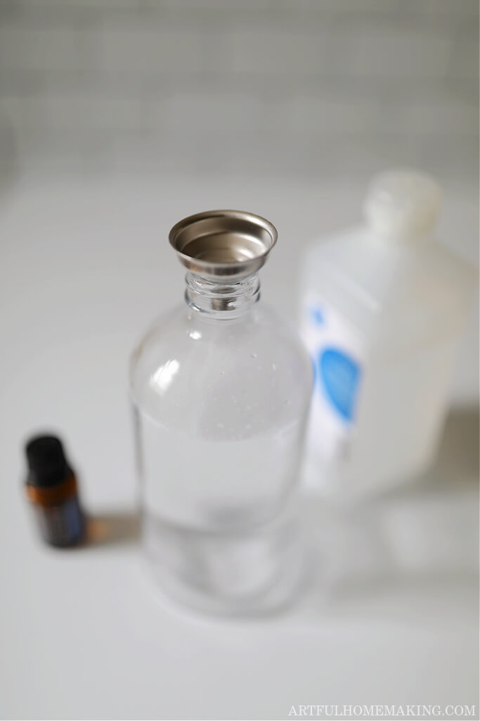 spray bottle with a metal funnel in the opening next to a bottle of rubbing alcohol and an essential oil bottle on a marble countertop