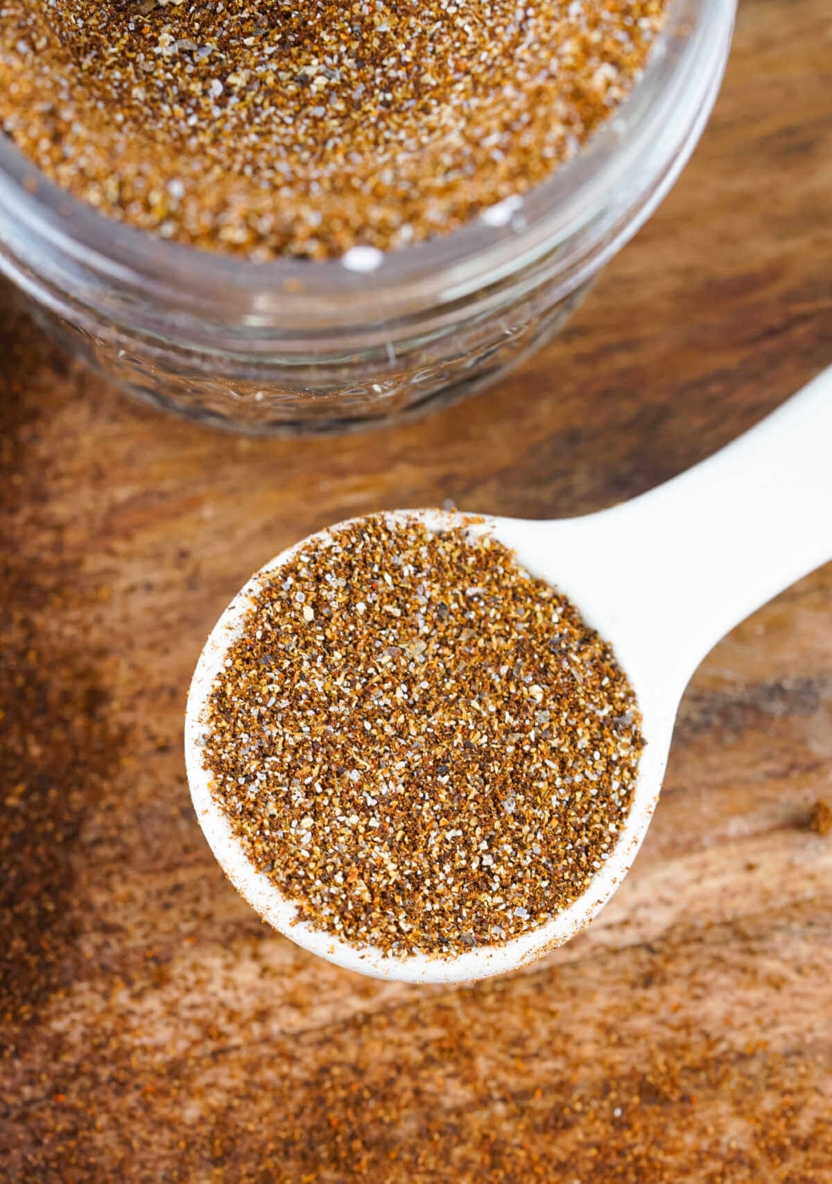 A measuring spoon filled with chili seasoning next to a jar of homemade chili seasoning mix.