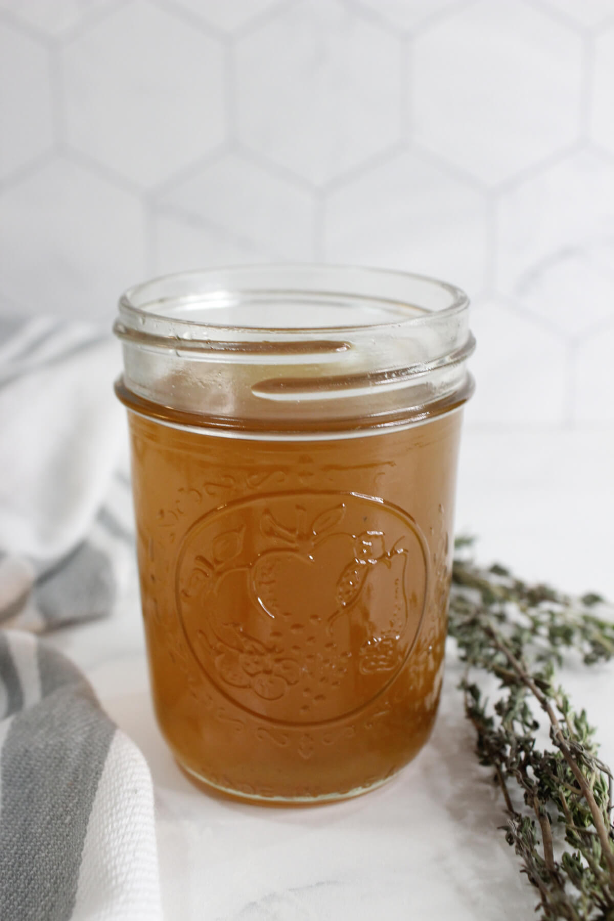 small mason jar sitting on a white countertop with homemade cough syrup next to a white towel with gray stripes and a sprig of fresh thyme