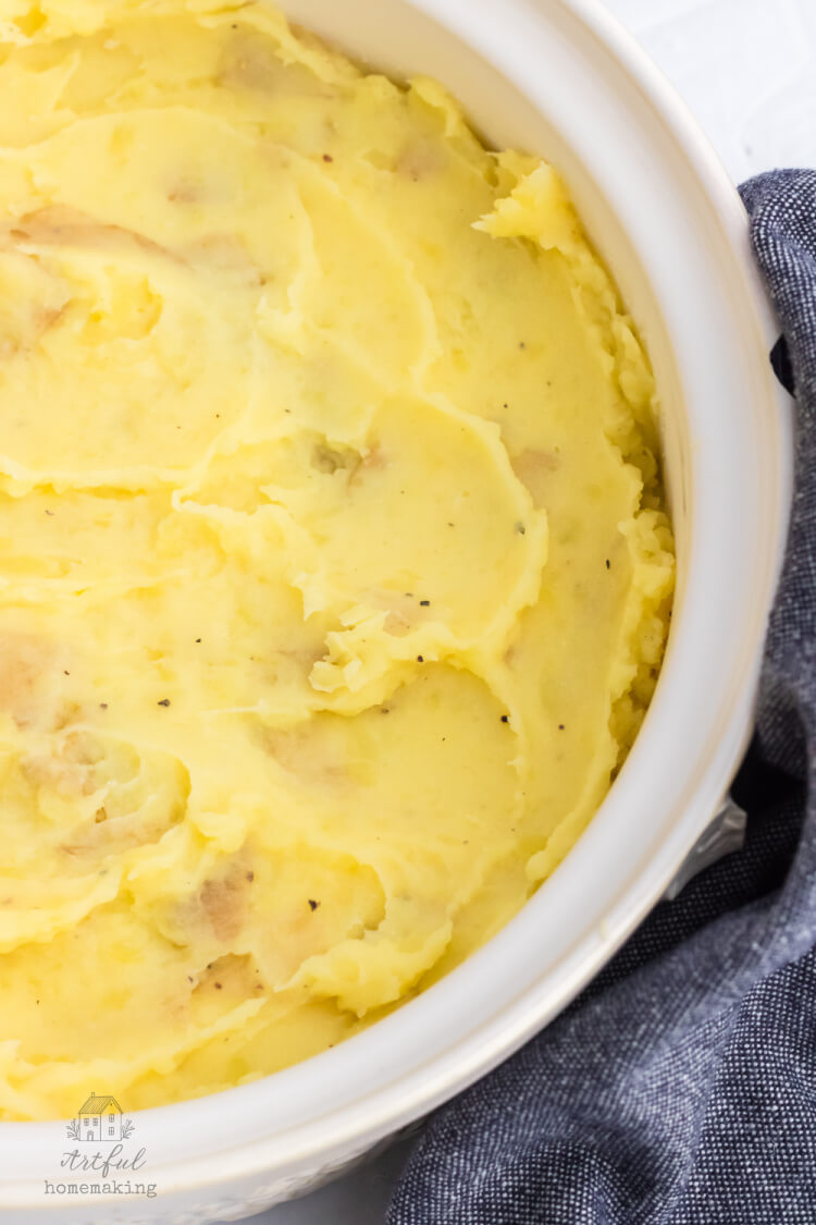 closeup of mashed potatoes in a white pot with a blue dish towel next to the pot