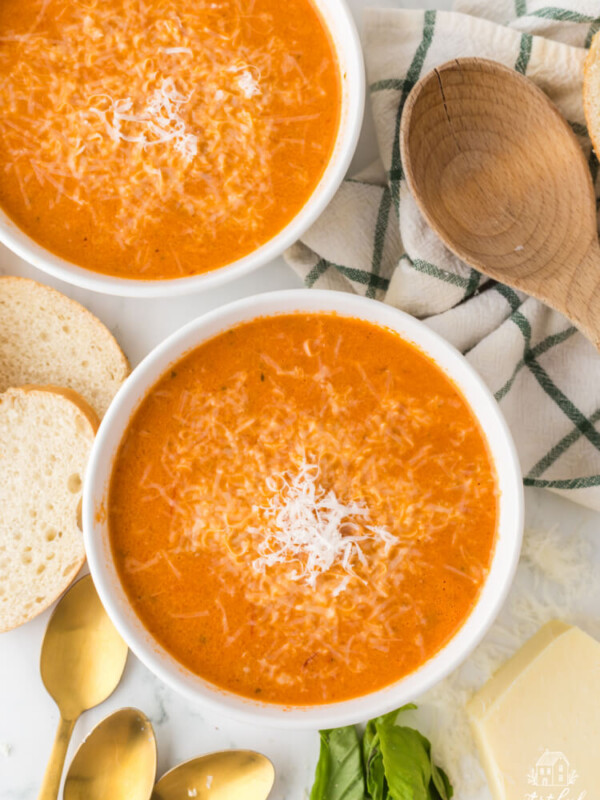 two white bowls of tomato soup on a marble countertop with sliced bread and parmesan cheese