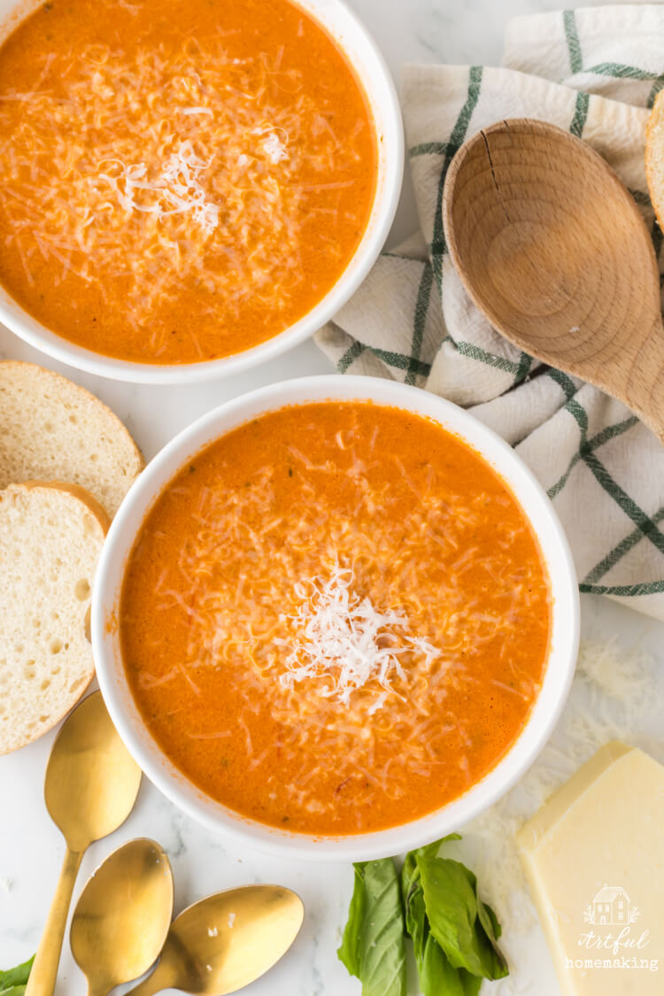 two white bowls of tomato soup on a marble countertop with sliced bread and parmesan cheese