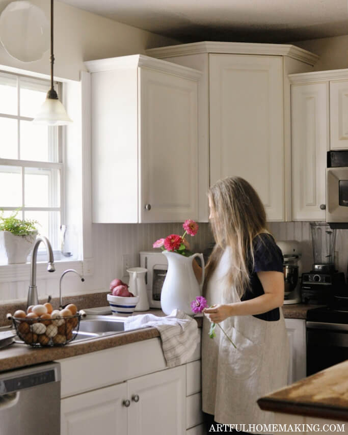 woman standing at the kitchen sink putting flowers in a white pitcher for decoration