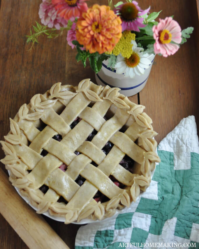 homemade pie with lattice top crust sitting on a table with a vase of flowers, a quilt, and a rolling pin