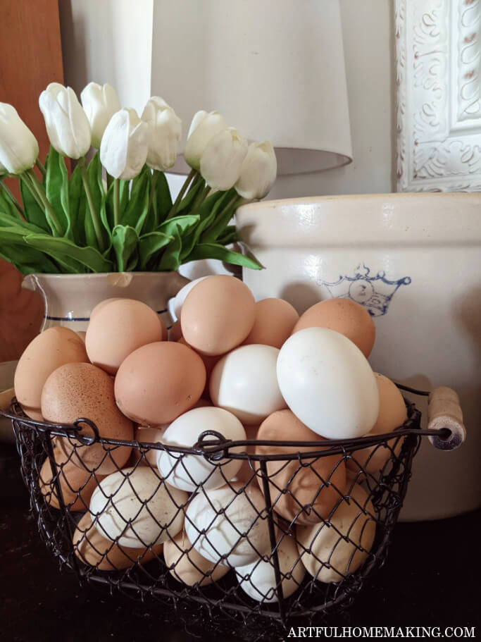 wire basket filled with white and brown chicken eggs in front of a pitcher filled with white tulips