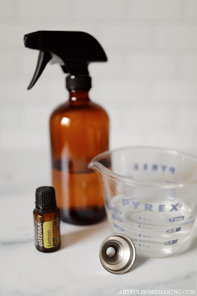 lemon essential oil, amber spray bottle, and glass measuring cup on a marble kitchen countertop