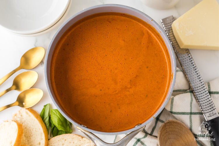 pot of roasted tomato soup next to three gold spoons, a wedge of parmesan cheese, and sliced bread