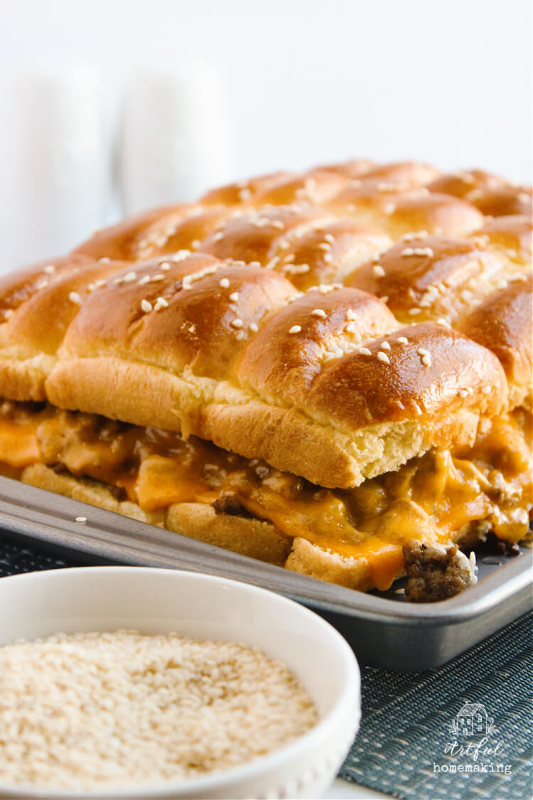 cheeseburger sliders on a metal baking sheet with sesame seeds in a white bowl in foreground