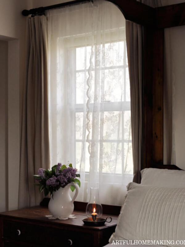 peaceful bedroom scene with lace curtains at the window, a ceramic pitcher with lilacs, and a lit candle next to a bed with white bedding