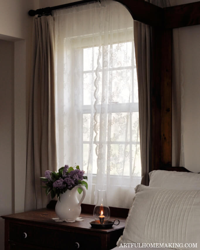 peaceful bedroom scene with lace curtains at the window, a ceramic pitcher with lilacs, and a lit candle next to a bed with white bedding