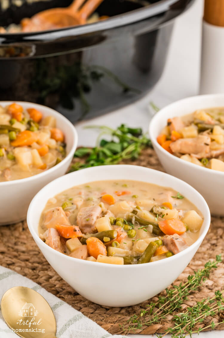 a black slow cooker insert in the background with three bowls of soup in the foreground