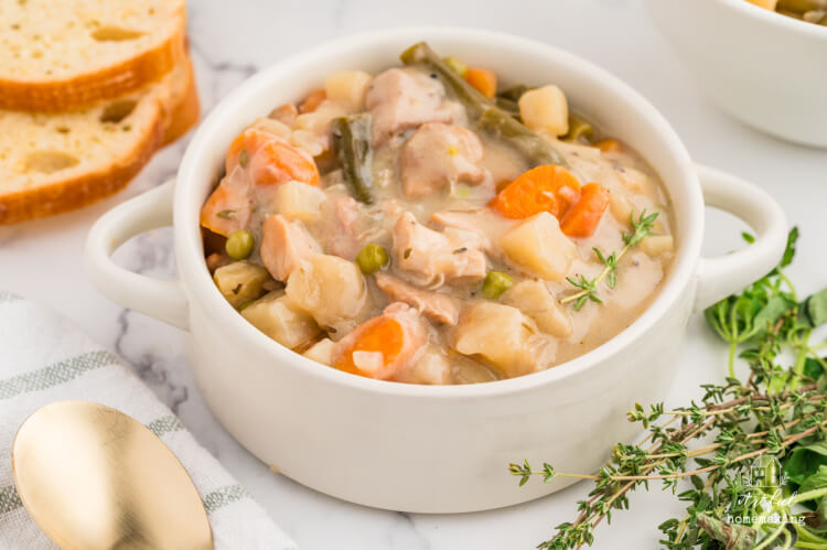 a white bowl of chicken stew with carrots, potatoes, and green beans sits on a marble surface with bread in the background and herbs in foreground