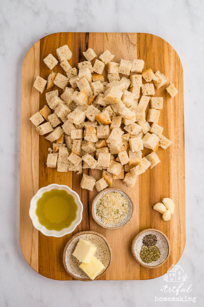 wooden cutting board with cubed sourdough bread, olive oil in a small bowl, butter in a small bowl, seasonings, and garlic cloves