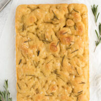 top down view of sourdough focaccia on a white counter with a bowl of olive oil and rosemary sprigs