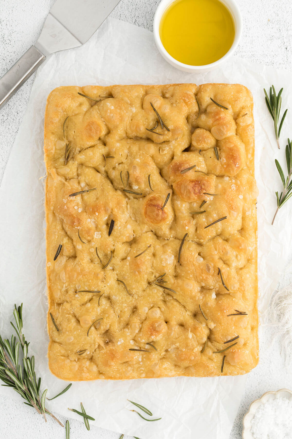 top down view of sourdough focaccia on a white counter with a bowl of olive oil and rosemary sprigs