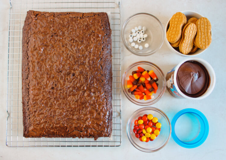 baked brownies on a cooling rack with candy in little glass bowls, a bowl of nutter butter cookies, and a container of chocolate frosting